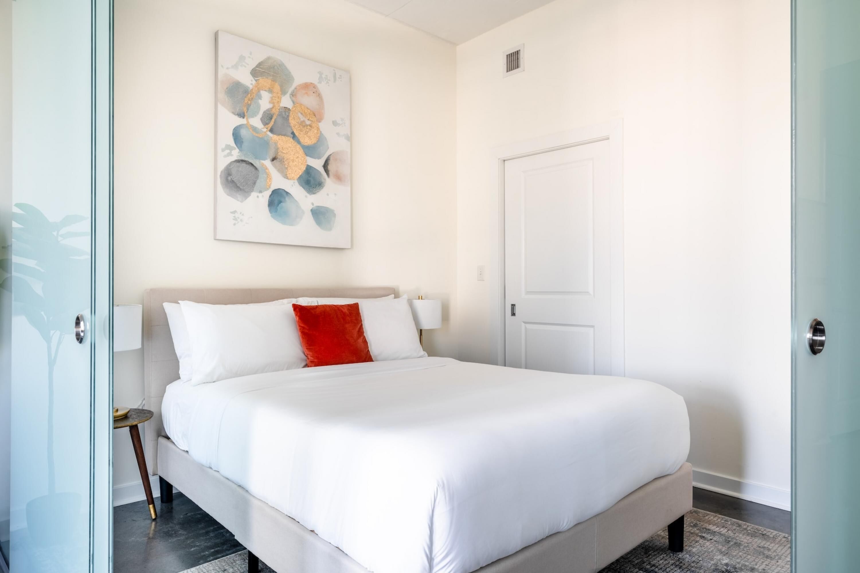 Een moderne slaapkamer met een groot bed, een nachtkastje, een abstract schilderij aan de muur en een deur.