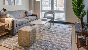 A modern living room with a sofa, coffee table, and ottoman, a ceiling fan, and a large window with blinds. - Kasa Family Friendly Westown 2BD Amenity Access Milwaukee (Milwaukee)