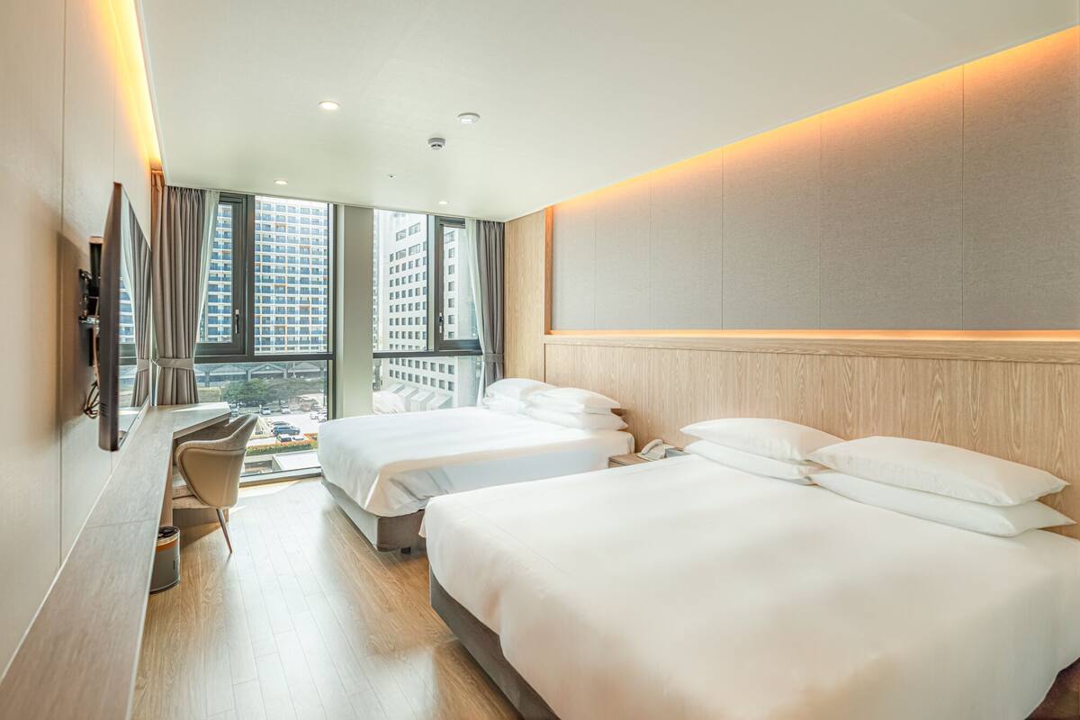 A hotel room with two beds, a desk, and a large window offering a city view.