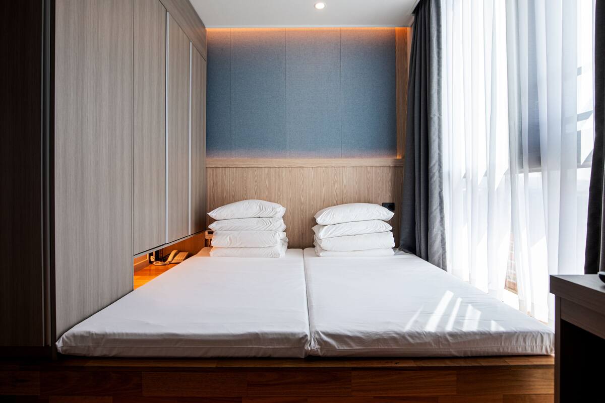 A double bed with white linens, a wooden headboard, and a light-colored wall.