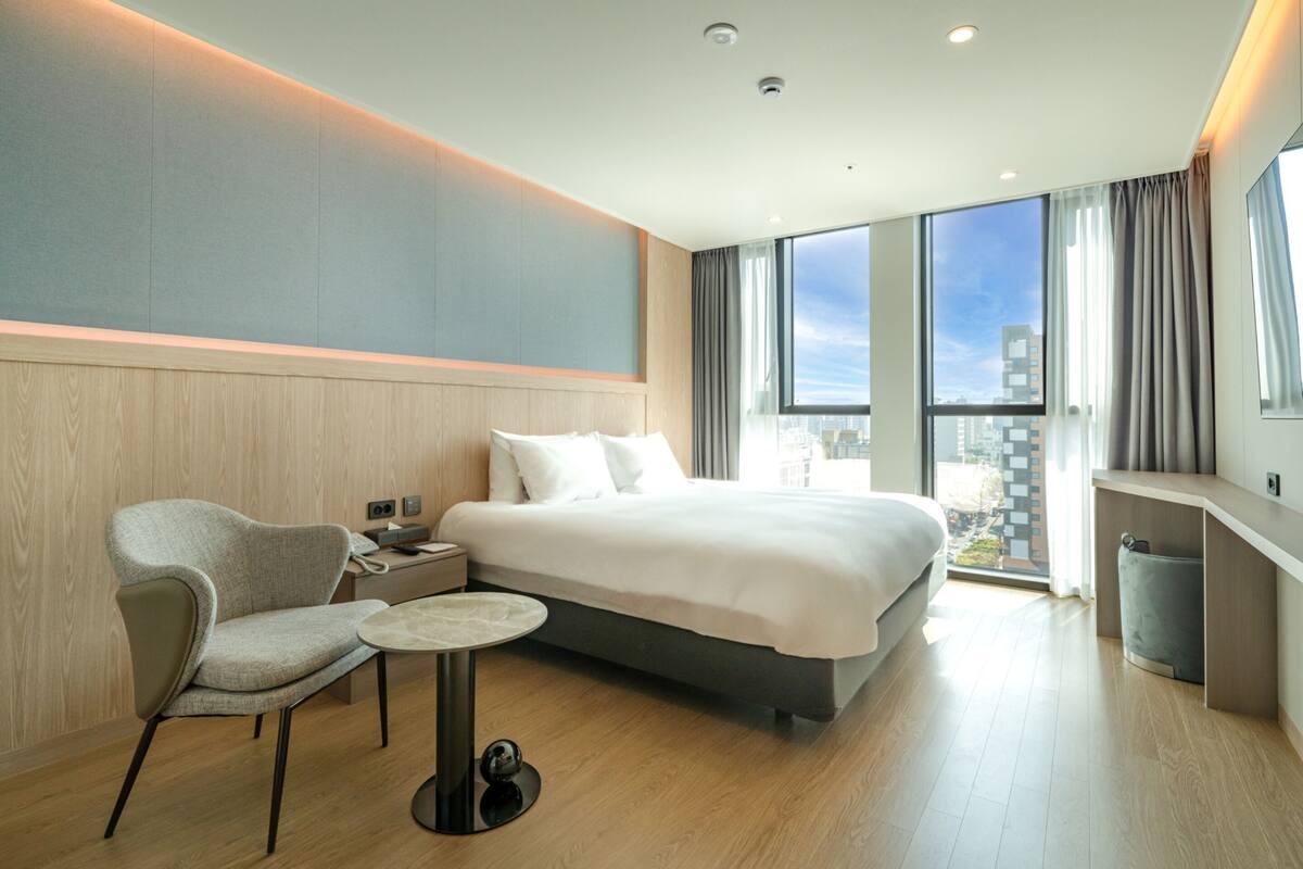 A modern hotel room with a large bed, a seating area with a chair and table, and a city view through large windows.