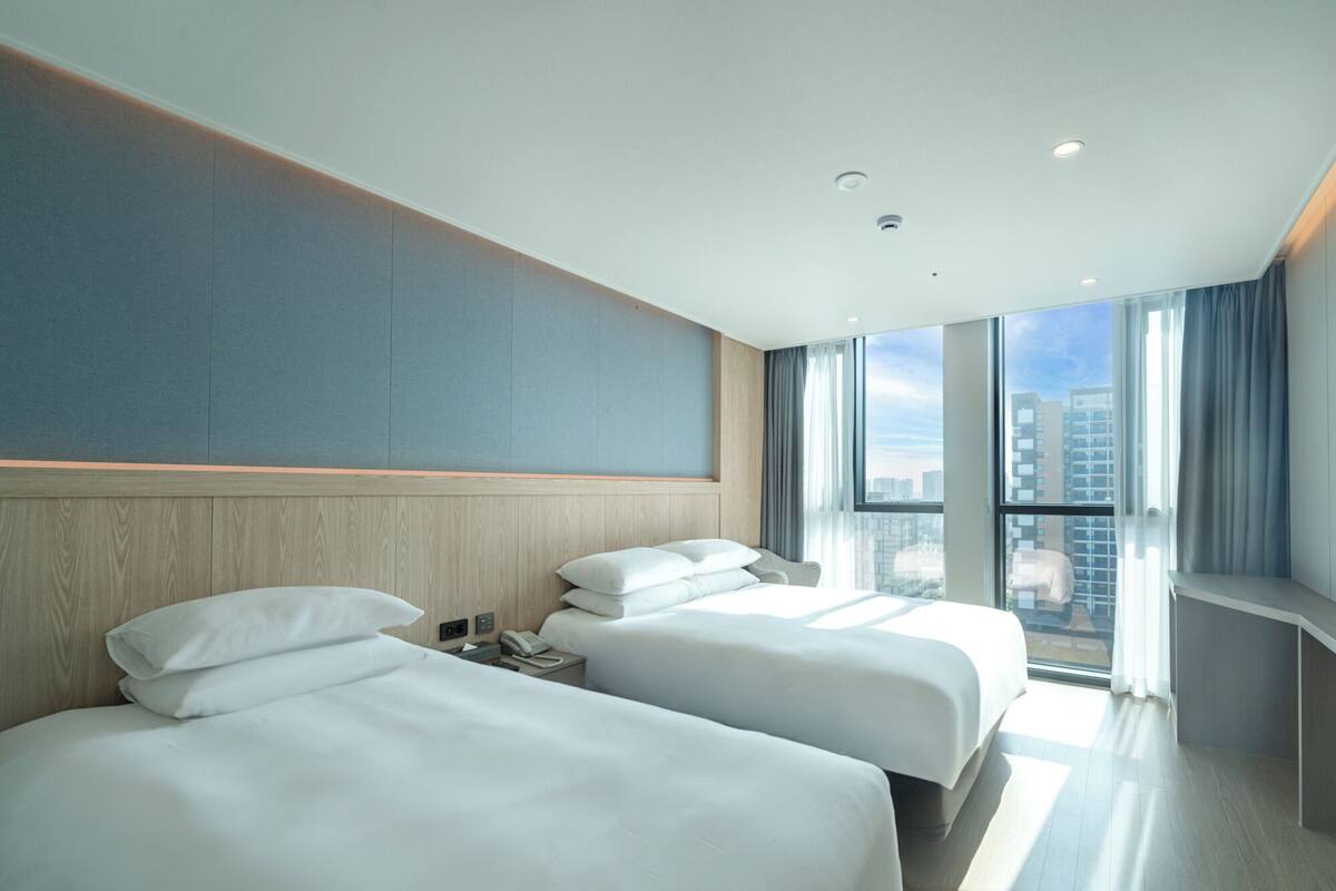 A modern hotel room with two beds, a large window with a city view, and a wooden headboard.