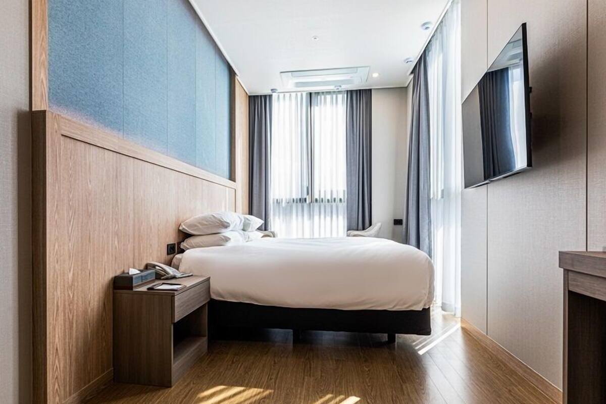 A modern hotel room with a large bed, wooden flooring, a bedside table with a phone, and a flat-screen TV mounted on the wall.