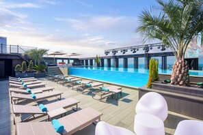 Ein Poolbereich auf dem Dach mit Liegestühlen und Sonnenschirmen, eine Bühne für Veranstaltungen und Blick auf die Skyline der Stadt.