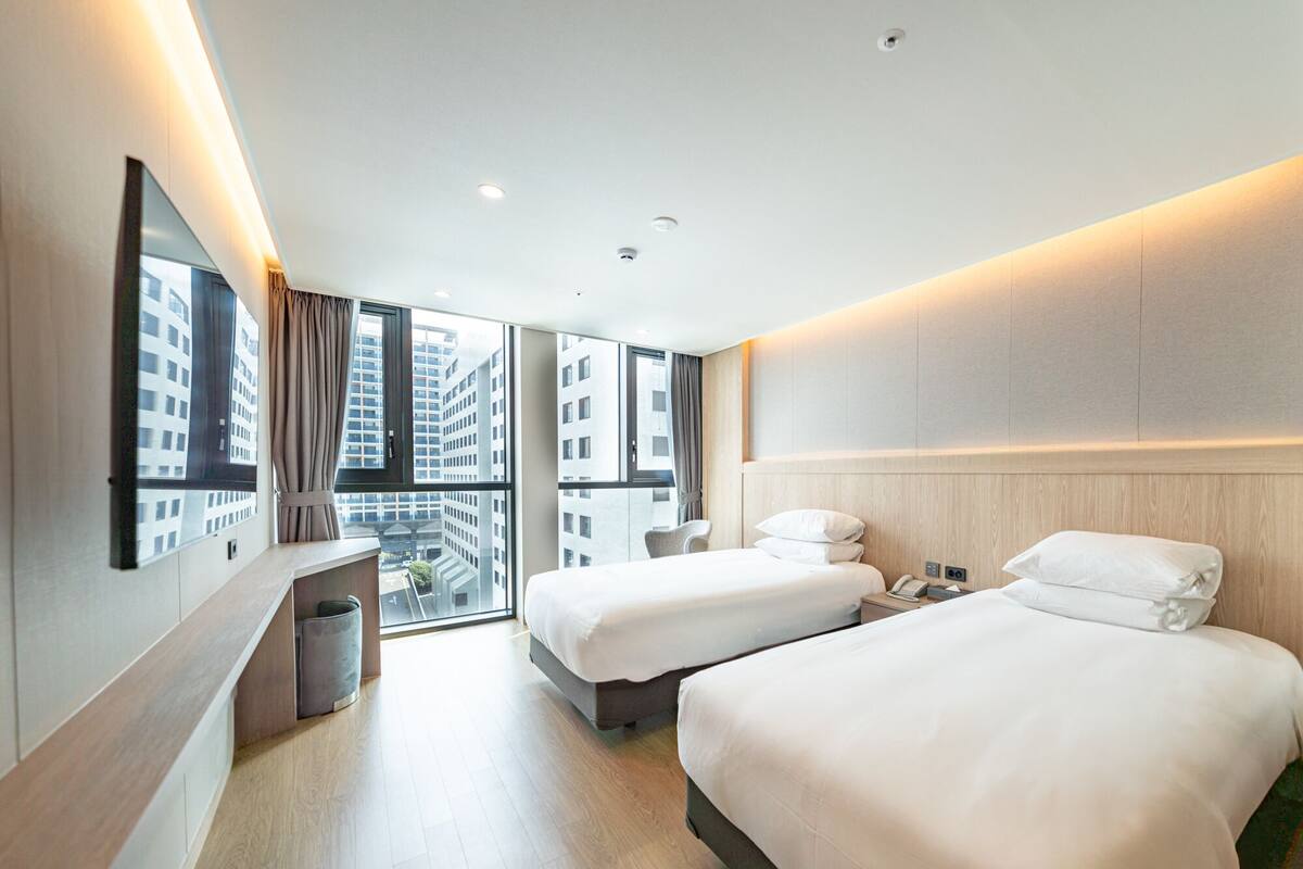 A modern hotel room with two beds, a desk, and a large window offering a city view.