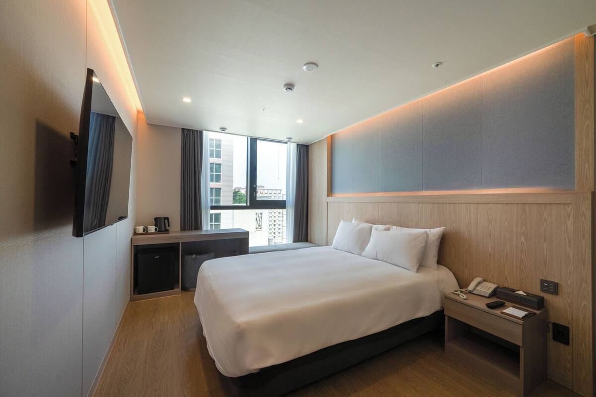 A modern hotel room with a large bed, a desk with a chair, a flat-screen TV mounted on the wall, and a window with a view of buildings.