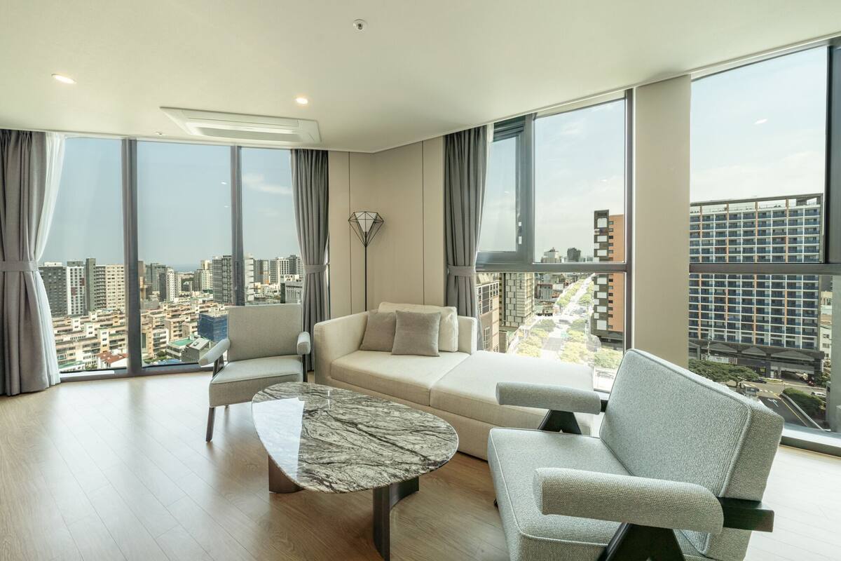 A modern living room with a sofa, armchairs, and a marble coffee table. Large windows offer a city view.