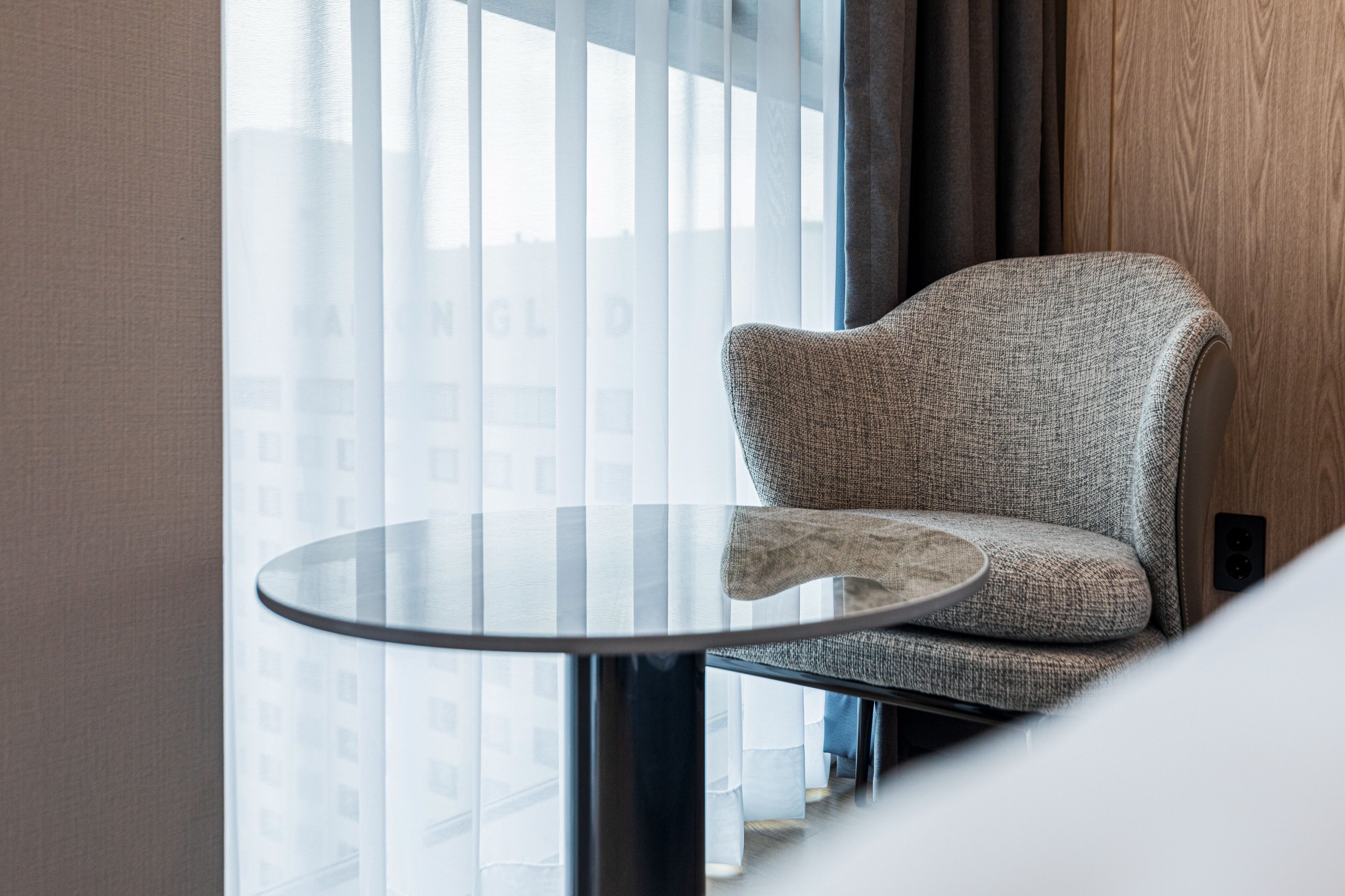 Quarto moderno com mesa de vidro redonda, cadeira estofada cinza e vista para edifícios através de cortinas leves.