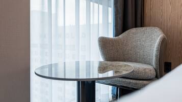 Quarto moderno com mesa de vidro redonda, cadeira estofada cinza e vista para edifícios através de cortinas leves.