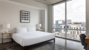 Una camera d'albergo moderna con un letto grande, due comodini con lampade, un quadro appeso alla parete e vista sulla città attraverso ampie finestre.