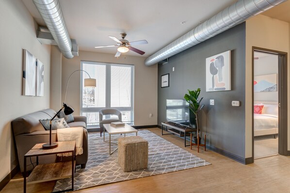 Un salon moderne comprenant un canapé, une table basse et un pouf, un ventilateur de plafond et une grande fenêtre avec des stores.