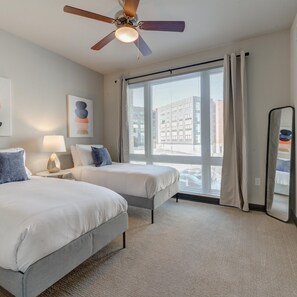 Une chambre d’hôtel avec deux lits, un ventilateur de plafond, une grande fenêtre avec des stores, un miroir et des œuvres d’art accrochées au mur.