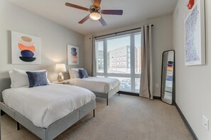 Une chambre d’hôtel avec deux lits, un ventilateur de plafond, une grande fenêtre avec des rideaux et des œuvres d’art accrochées aux murs.