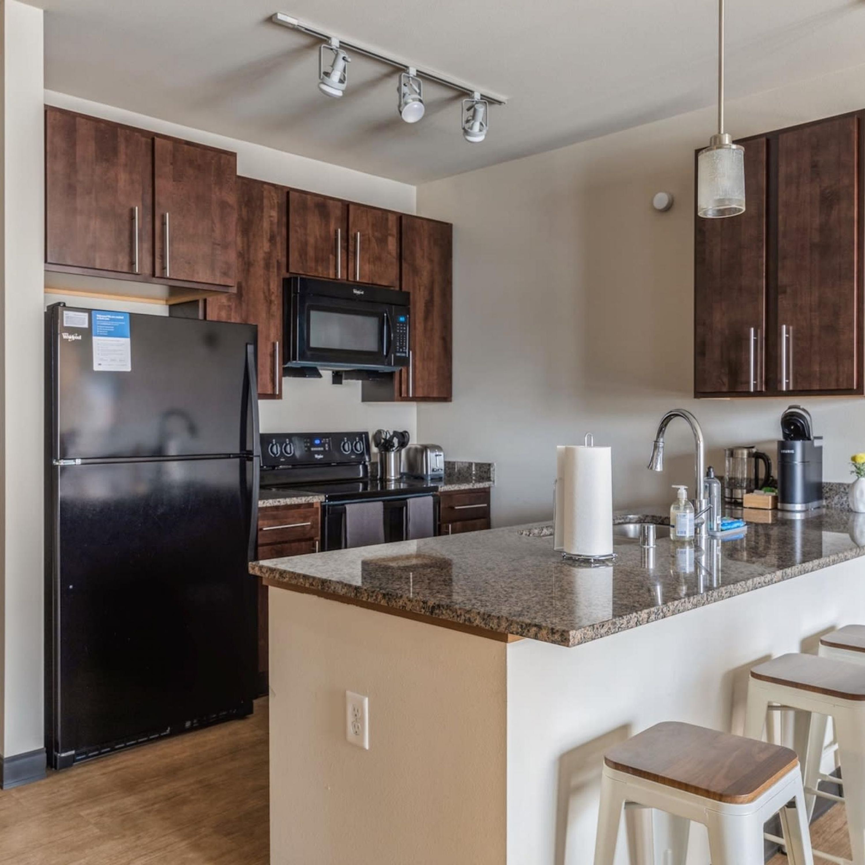 A modern kitchen with dark wooden cabinets, a black refrigerator, a microwave, a stove, and a granite countertop. There are three white bar stools and a hanging light fixture.