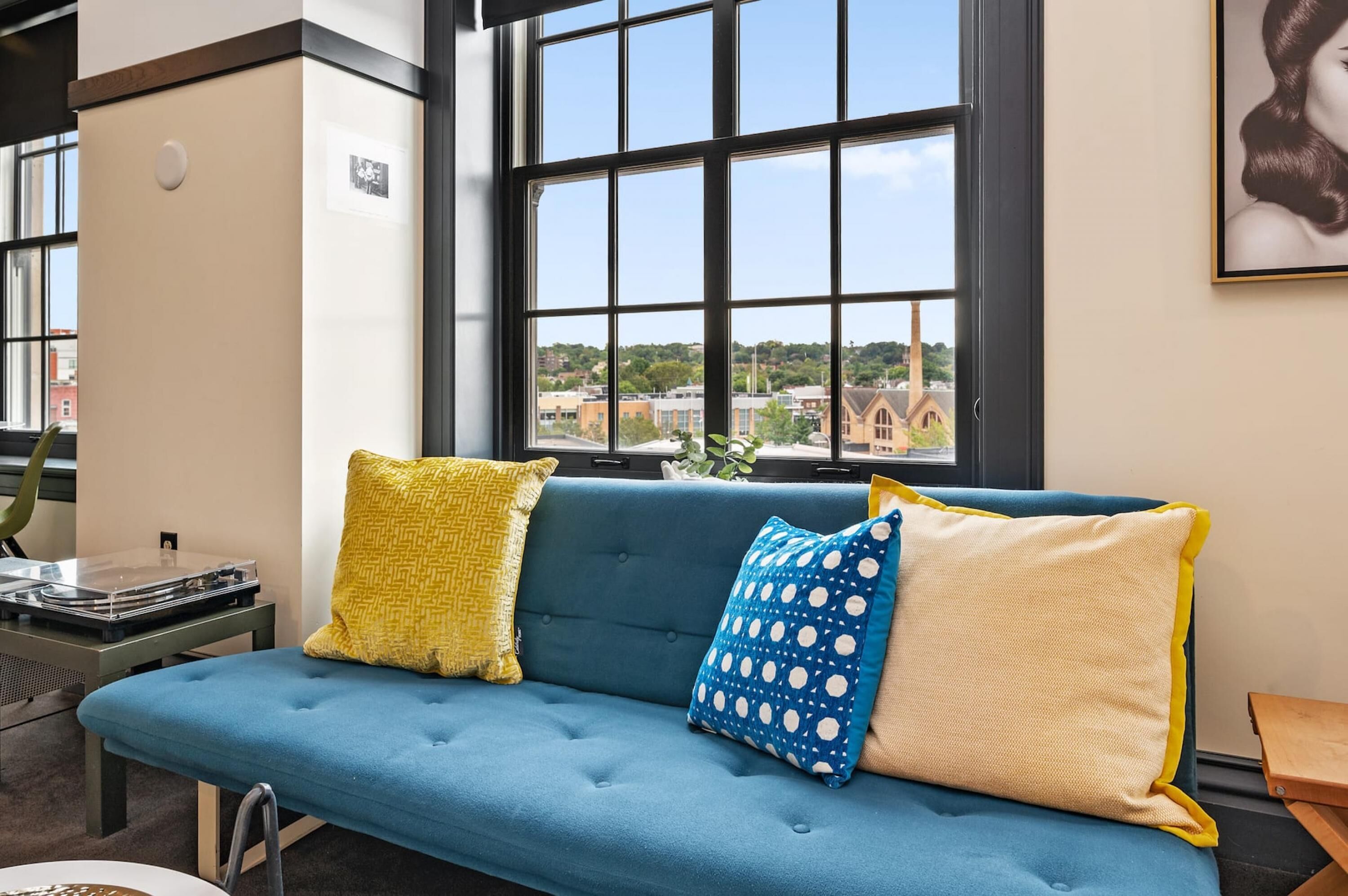 A blue sofa with patterned cushions, a turntable, and a window with a view of buildings and greenery.