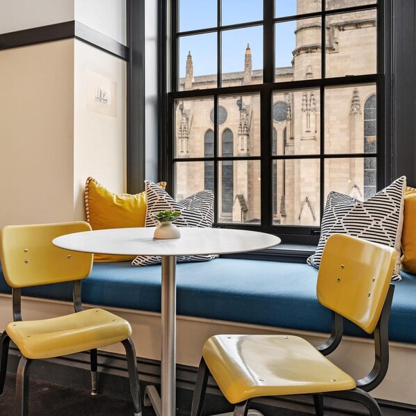 A corner seating area with a round table and two yellow chairs, a blue sofa with patterned cushions, and a view of historic architecture through large windows.