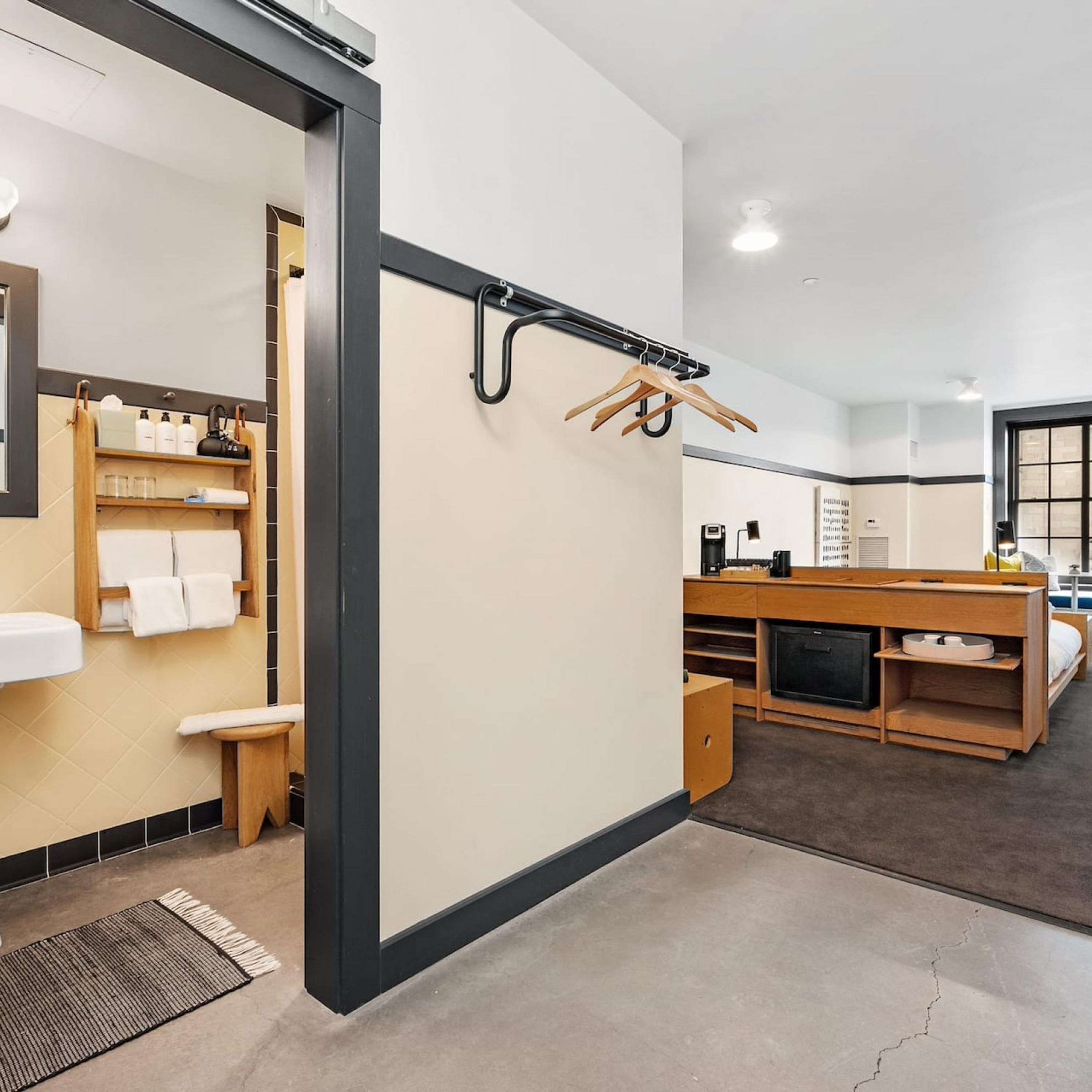 A modern hotel room with a bathroom, a wooden vanity, a bed, and a small seating area.