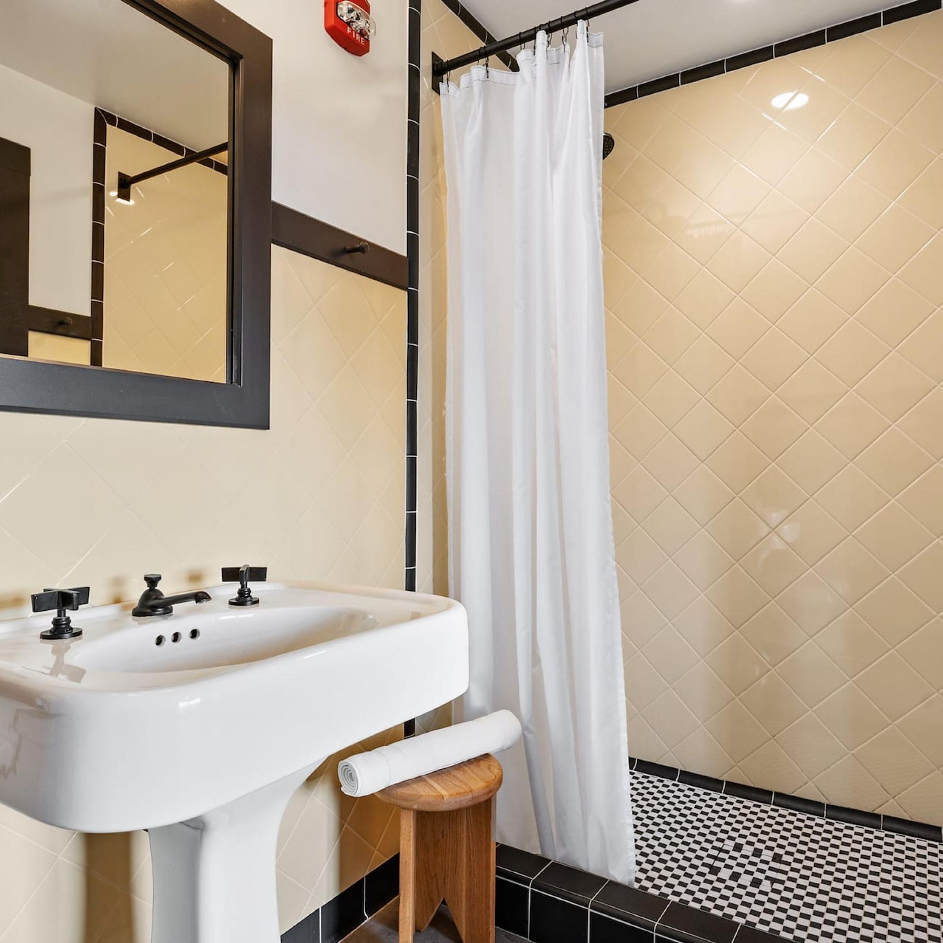 A bathroom with a white sink, a wooden stool, a shower curtain, and a fire alarm on the wall.