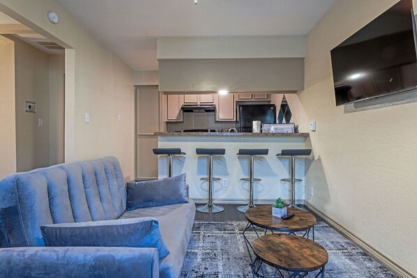 Uma sala de estar moderna com um sofá azul, uma mesinha de centro e uma área de cozinha com um banquinho e uma televisão.