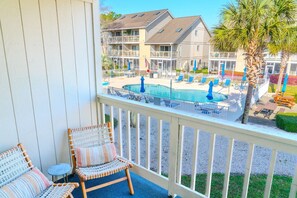A balcony with wicker chairs, a pool area with lounge chairs and umbrellas, and a view of multi-story buildings. - Golf Colony at Deerfield 19-D (Myrtle Beach)
