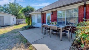 A residential house with a patio area featuring a table and chairs, a covered outdoor seating area, and a visible garage. - Enjoy a nice home with a beautiful garden.   (Fort worth)