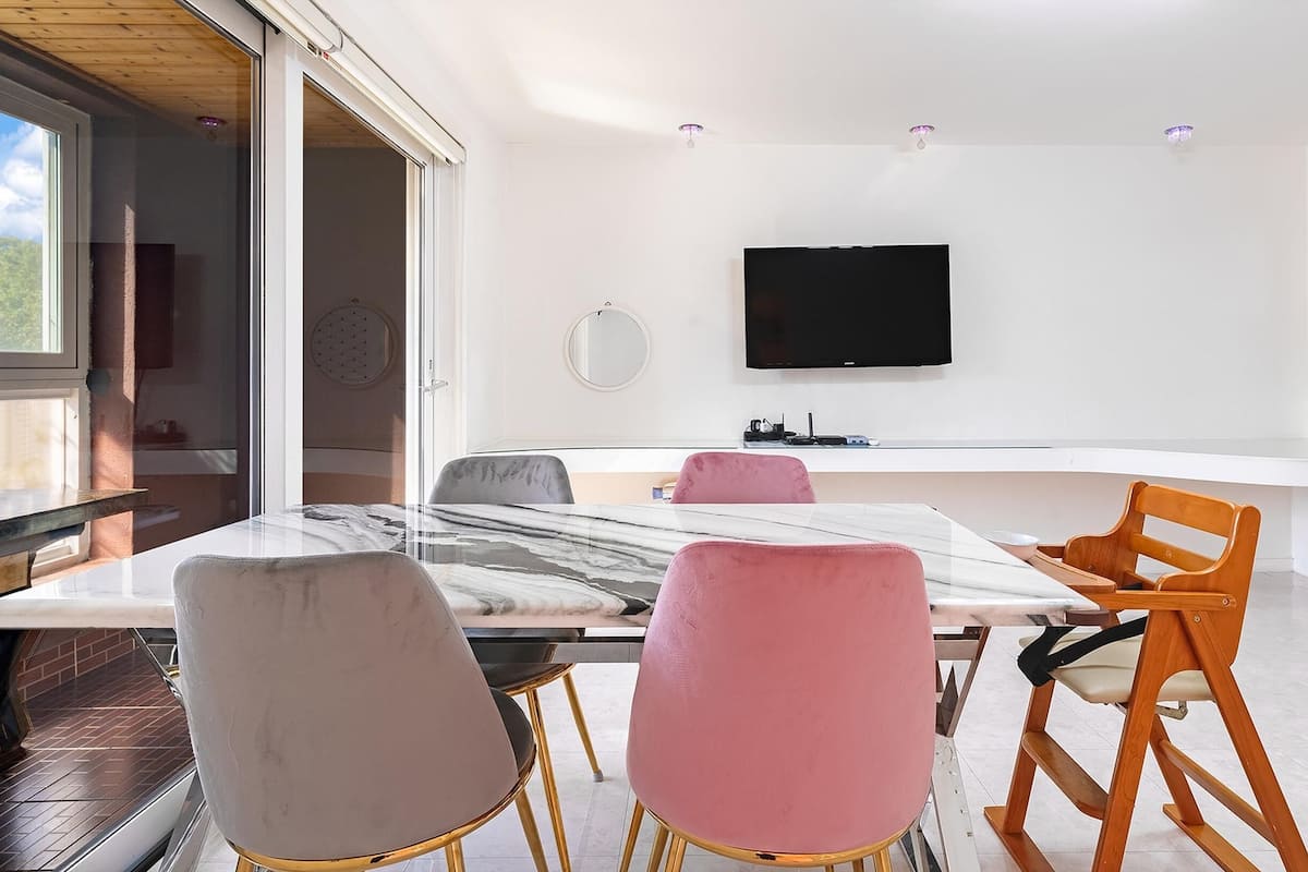 A modern dining area with a marble table, a flat-screen TV mounted on the wall, and a large window providing natural light.