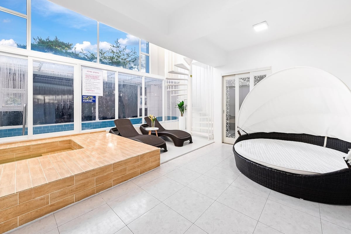 A modern indoor pool area with a wooden bench, lounge chairs, and a large wicker chair with a canopy.