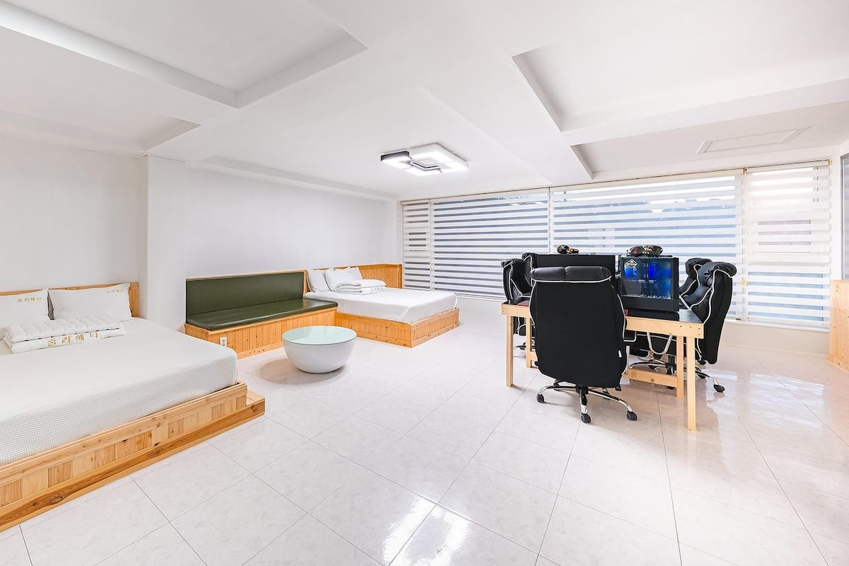 A modern hotel room with a wooden bed, a green sofa, a small round table, a desk with a computer, and a large window with blinds.