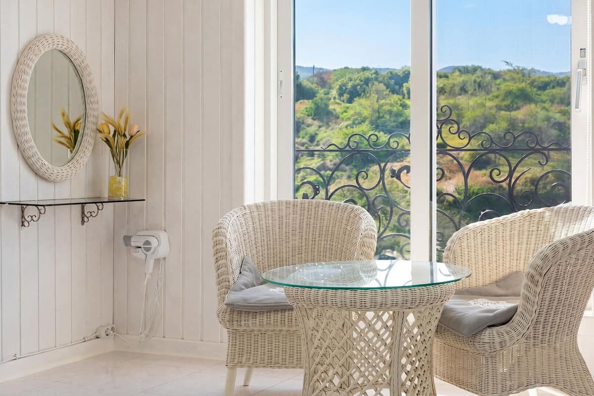 A balcony with wicker chairs, a glass table, a white wicker side table, a mirror, and a vase with flowers.