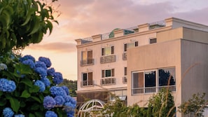 A multi-story building with balconies, large windows, and a garden with blue flowers.