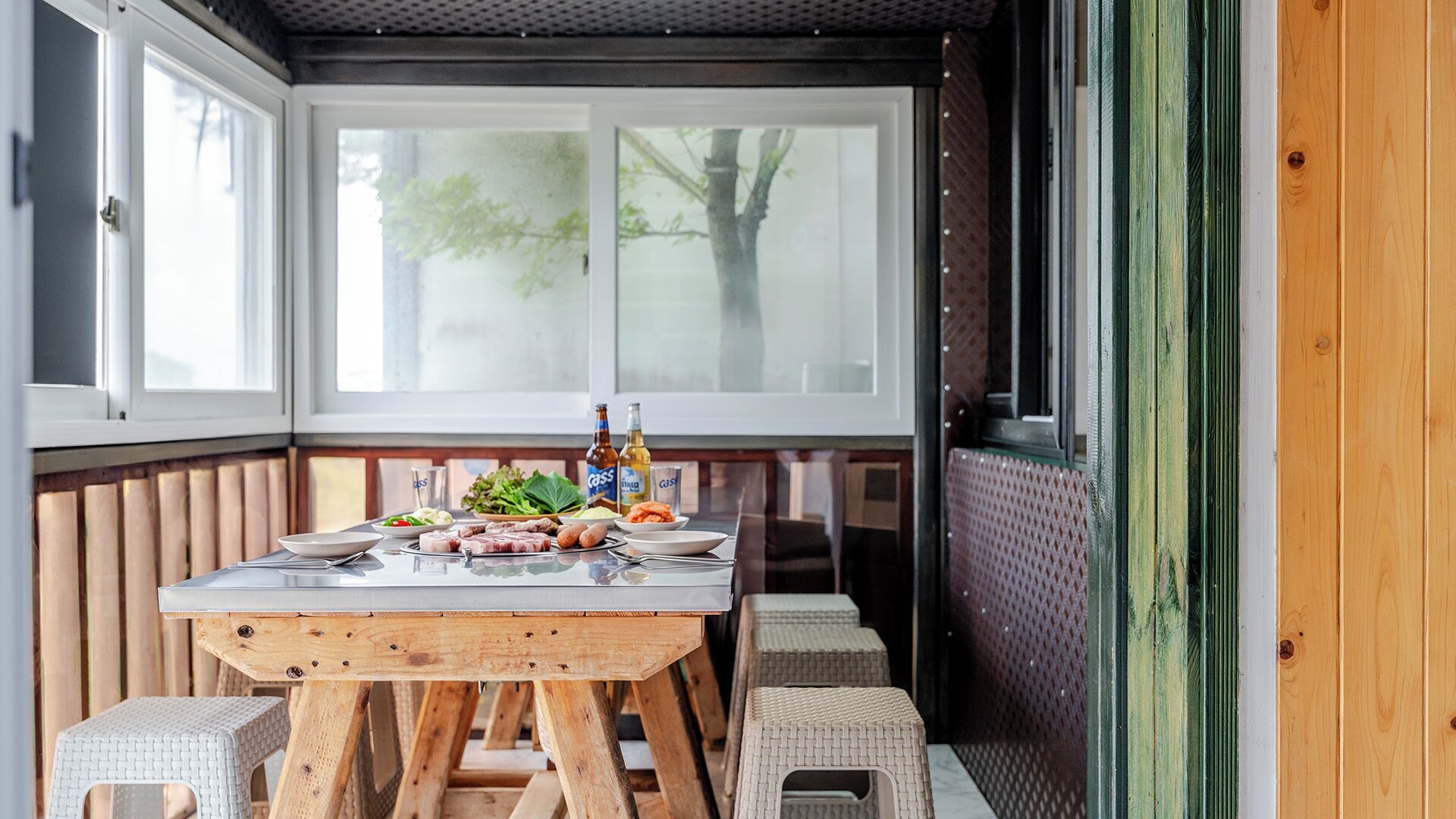 Un comedor con una mesa de madera dispuesta para una comida, rodeado de sillas de mimbre, y vista a árboles a través de amplios ventanales.