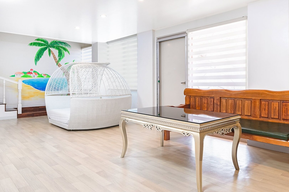 A room with a wooden bench, a glass-top table, a white wicker chair, and a wall mural.