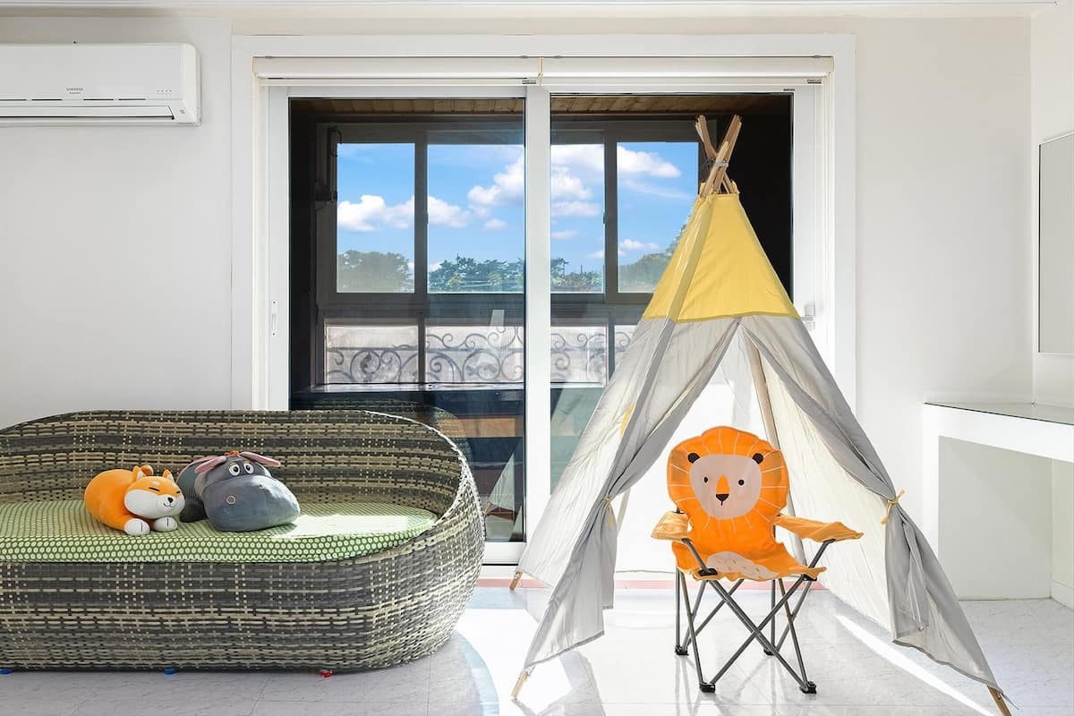 A room with a wicker sofa, a children's play tent, and a large window with a view of greenery.