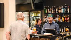 A bar area with a bartender and a customer. The bar has a variety of liquor bottles and a cash register. - Holbrook Hotel (Holbrook)
