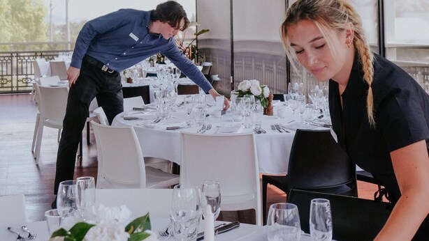 A restaurant setting with tables set for dining, a waiter attending to a table, and a woman seated at a table. - Holbrook Hotel (Holbrook)
