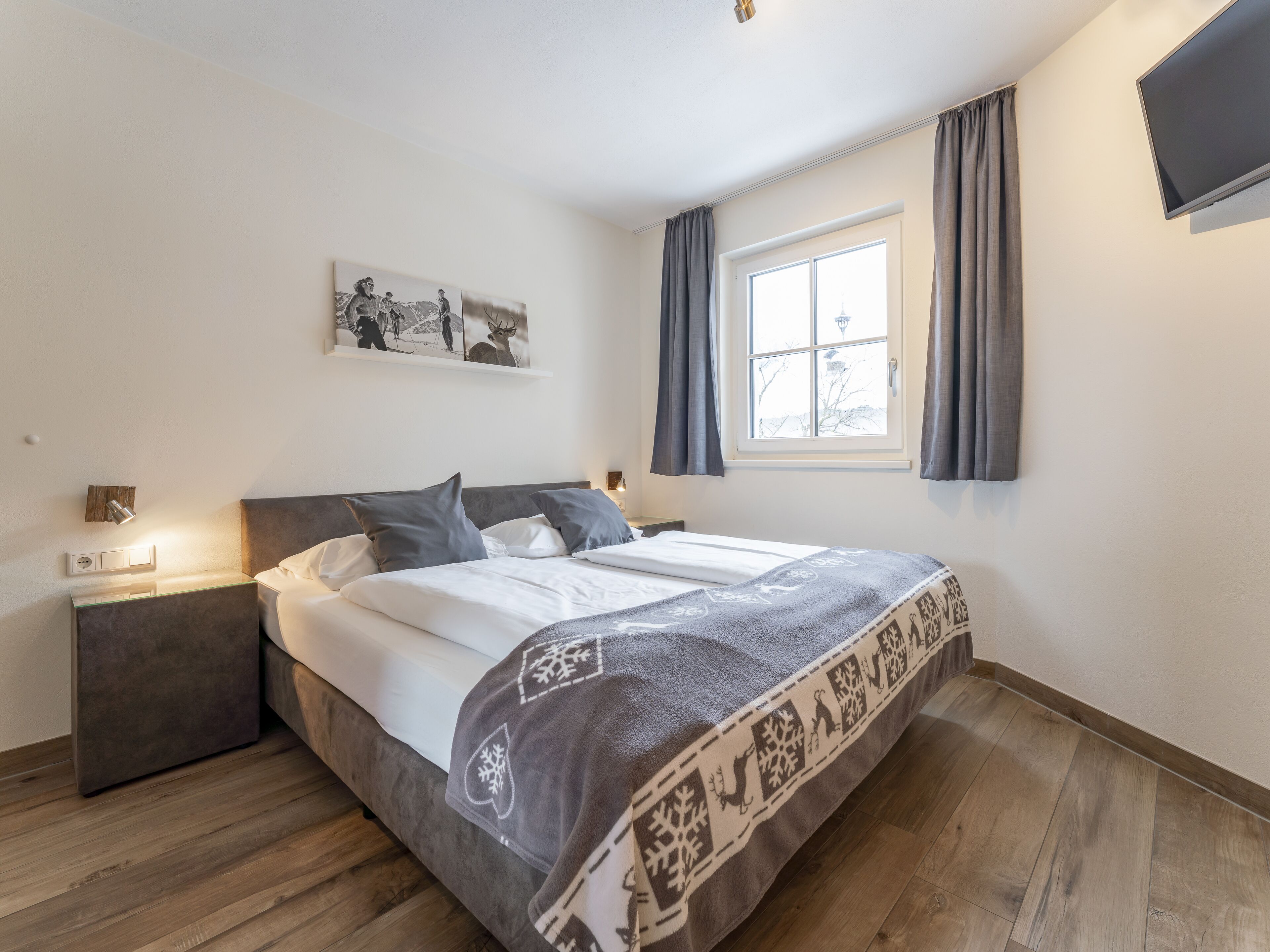 A bedroom with a bed, a nightstand, a wall-mounted TV, a window with curtains, and a painting on the wall.