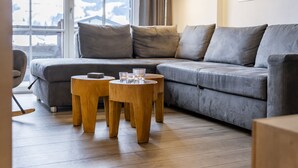 Ein modernes Wohnzimmer mit einem grauen Sofa, zwei Holznachttischen und einem großen Fenster mit Blick auf die Berge.