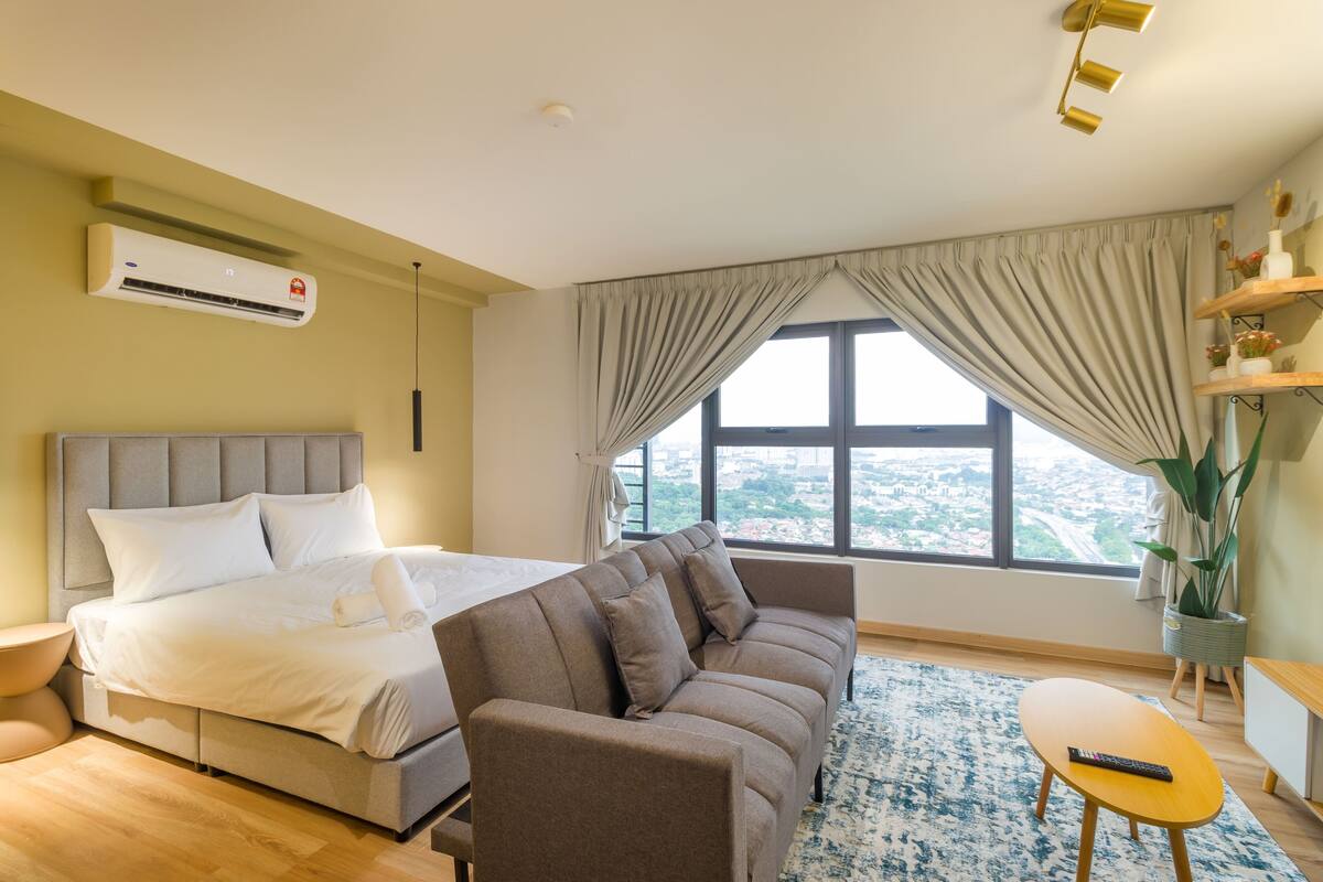 A modern hotel room with a large bed, a sofa, a small table, and a view of the city through the window.