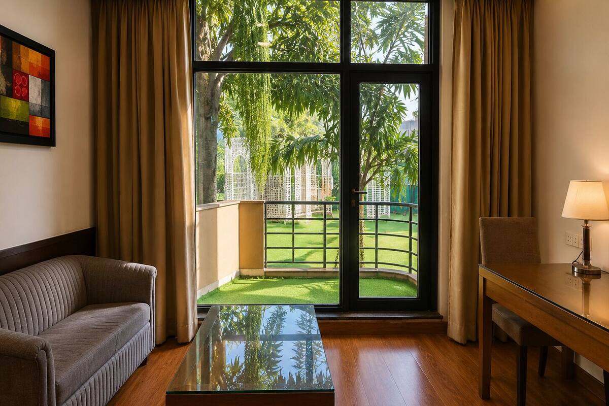 A room with a sofa, a glass-top table, a lamp, and a view of a garden through large windows.