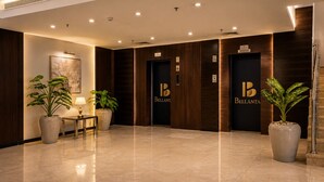 Eine Hotelhalle mit poliertem Marmorboden, zwei große Kübelpflanzen, ein Empfangstresen und zwei Aufzüge mit dem Hotel-Logo.