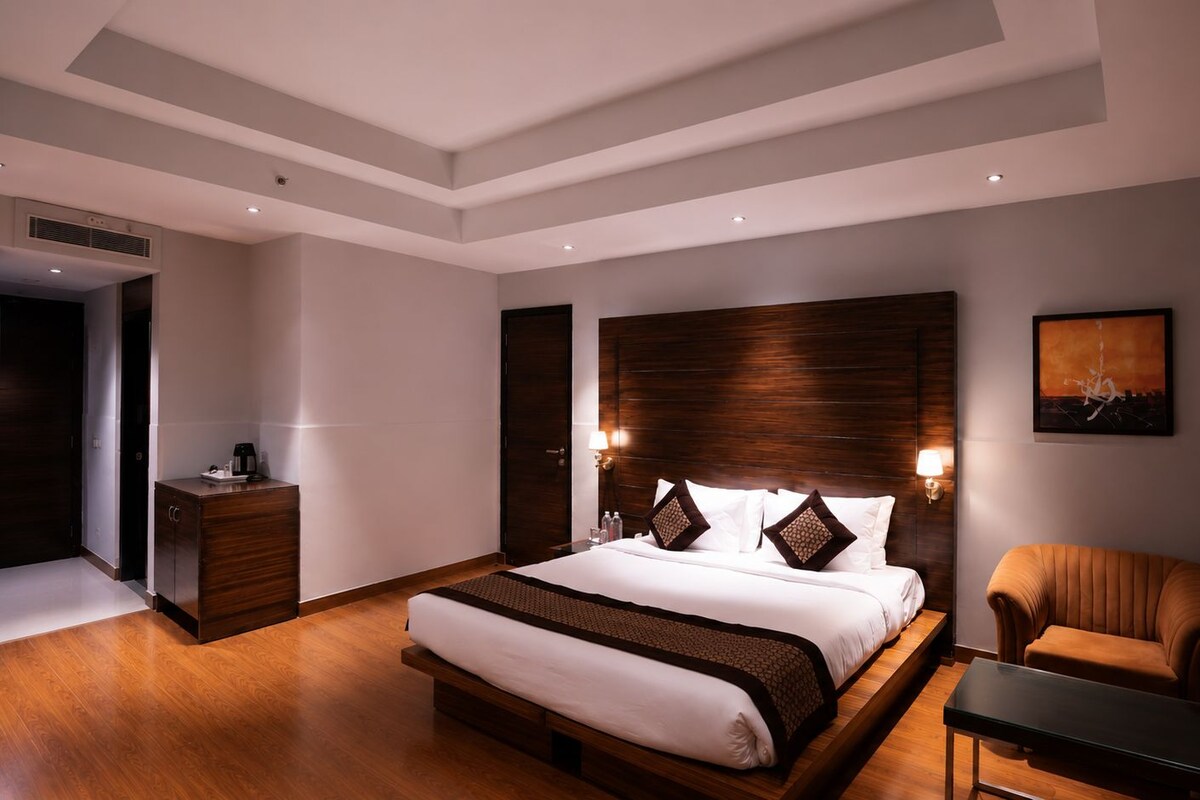 A modern hotel room with a large bed, a wooden headboard, a bedside table, a chair, and a painting on the wall.