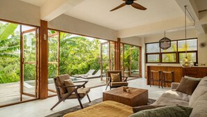 A spacious living area with a large glass door leading to a deck, a ceiling fan, and a bar area with stools. - Nowhere Villa (Pecatu)