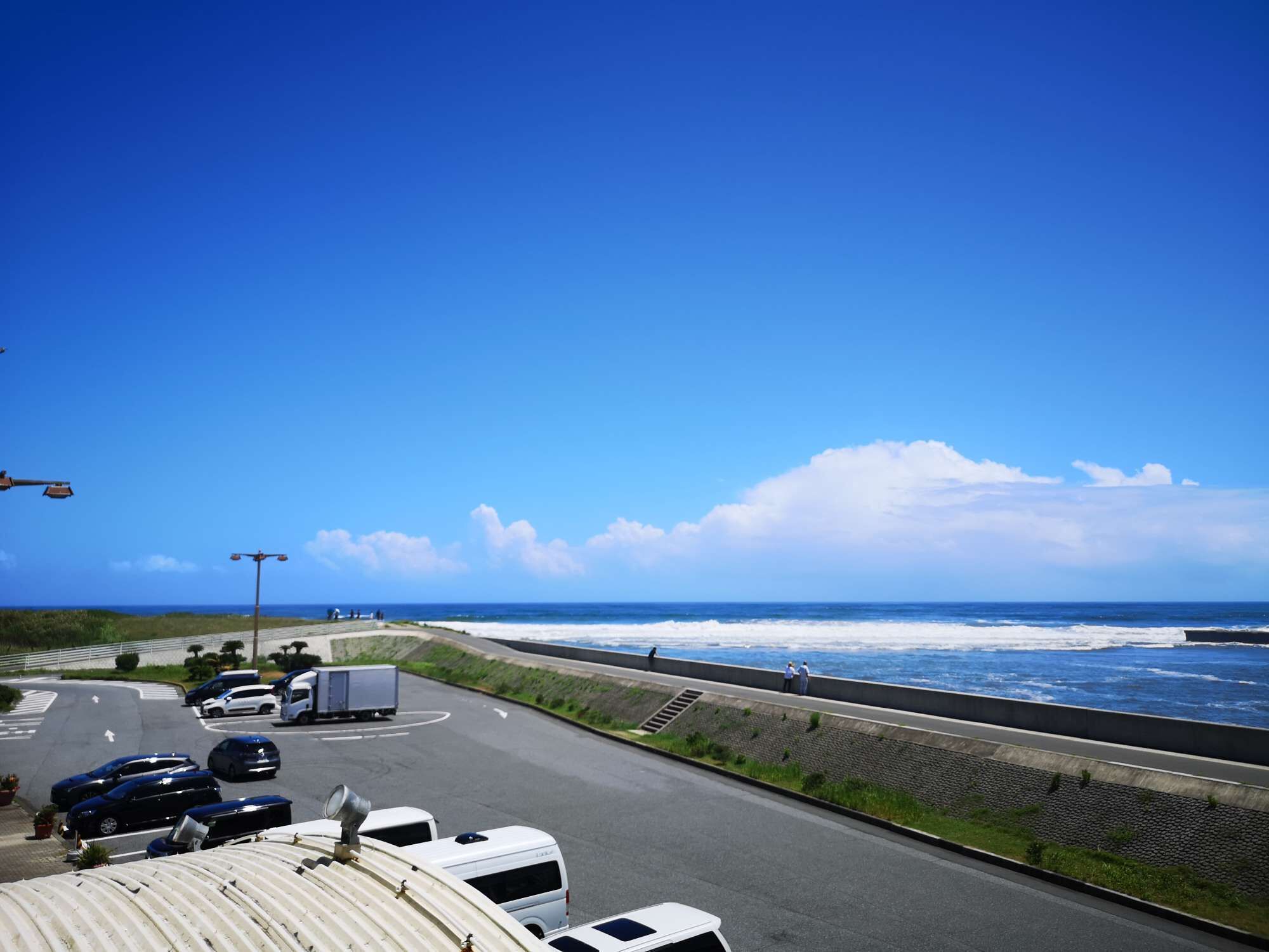 A coastal road with a view of the ocean, a parking area with cars and a bus, and a pedestrian walkway along the sea.