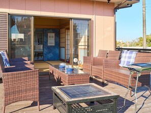 Een terras buiten met rieten meubels en een tafel, een schuifdeur die leidt naar een binnenruimte, en uitzicht op de omgeving.