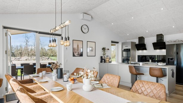 Una cocina moderna con electrodomésticos de acero inoxidable, un comedor con una larga mesa de madera y sillas acolchadas, y vistas al exterior a través de amplias ventanas.