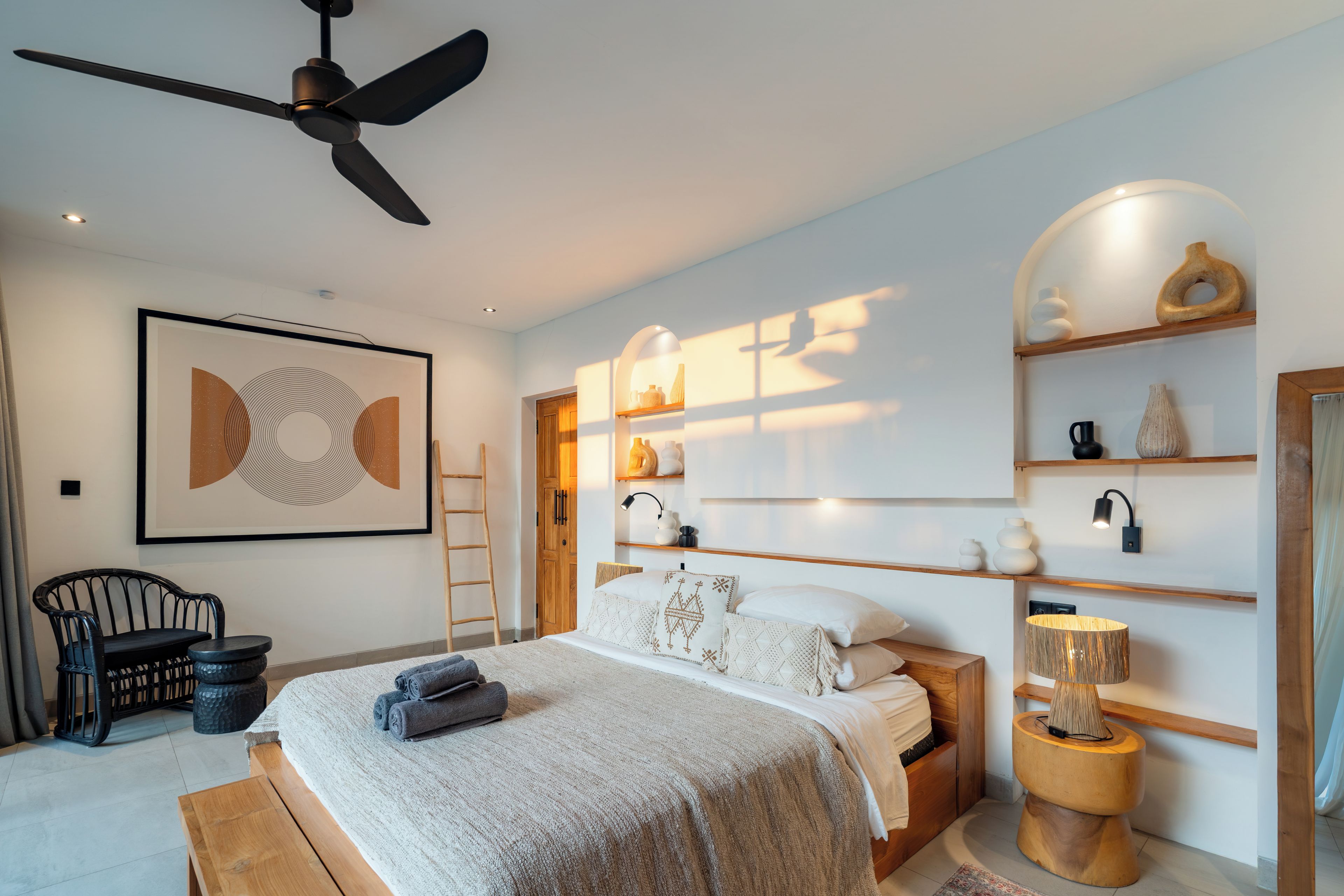 A modern bedroom with a bed, a ceiling fan, a framed artwork, a wooden ladder, and a bedside table with decorative items.