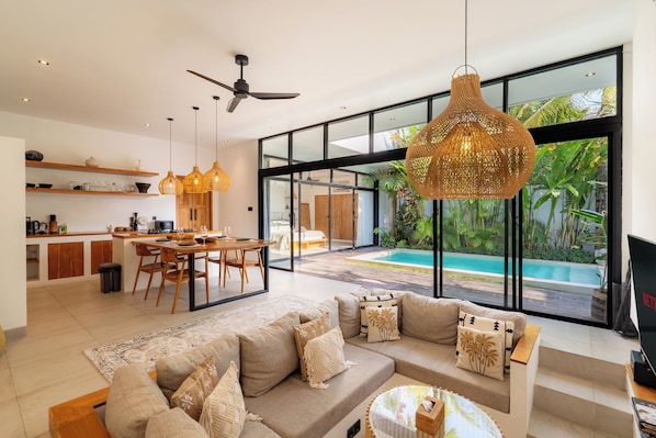 A modern living space with a dining area, kitchen, and a large glass door leading to a pool. - Jardin Villa by Betterplace (Pecatu)