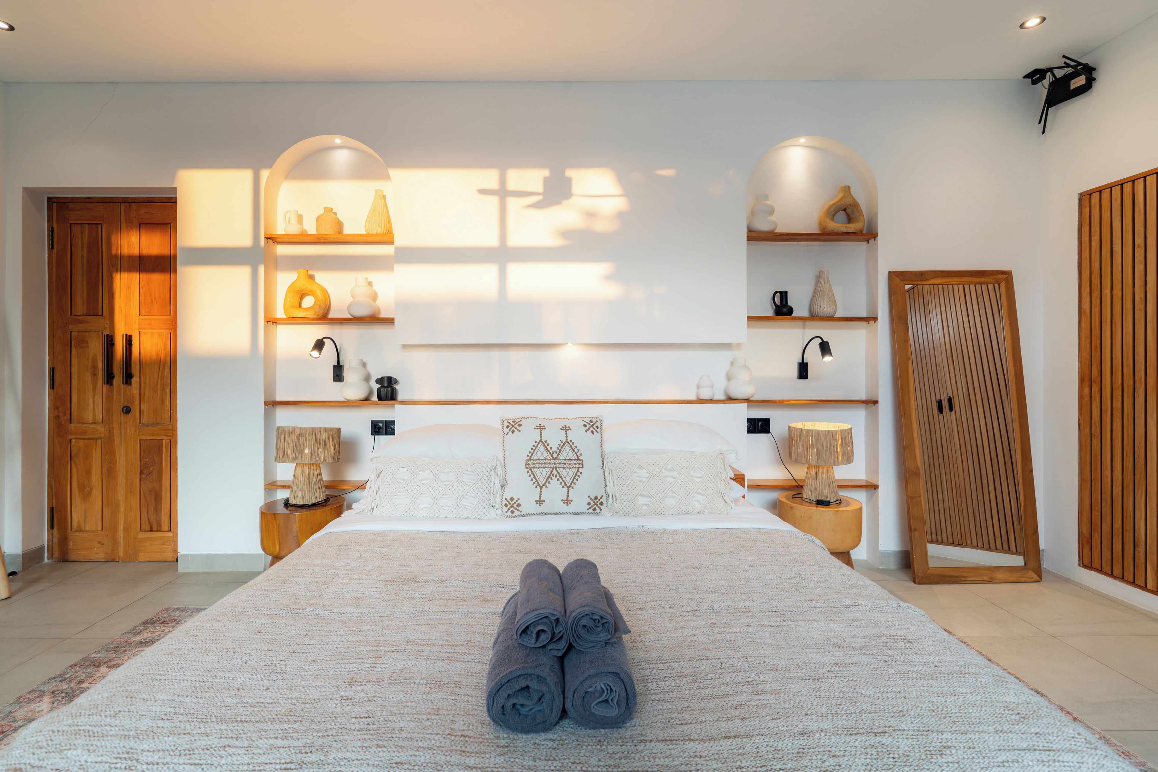 Un dormitorio moderno con una cama grande, dos mesitas de noche con lámparas, un armario y estantes con objetos decorativos.