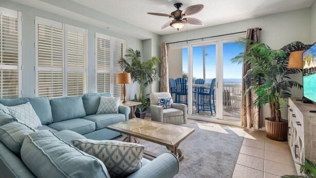 A spacious living room with a large sofa, a coffee table, and a ceiling fan. There are several chairs and a TV. The room has large windows with shutters and a view of the ocean. - Seychelles 101 (Panama City Beach)