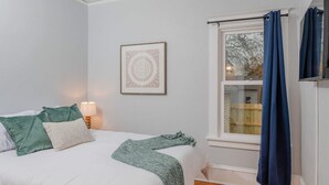 Un lit bien fait, avec une literie blanche et verte, une œuvre d’art encadrée au mur et une fenêtre avec des rideaux.
